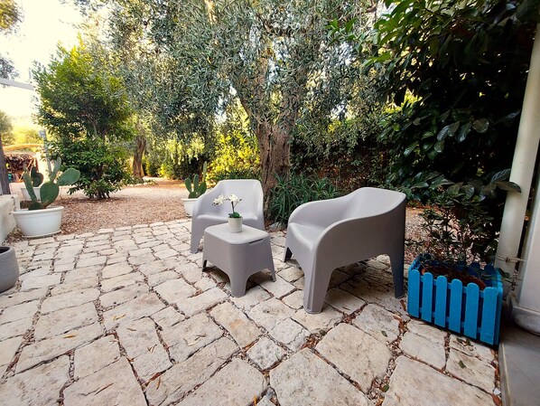 A patio with stone flooring, two white plastic chairs, a small table, and potted plants.