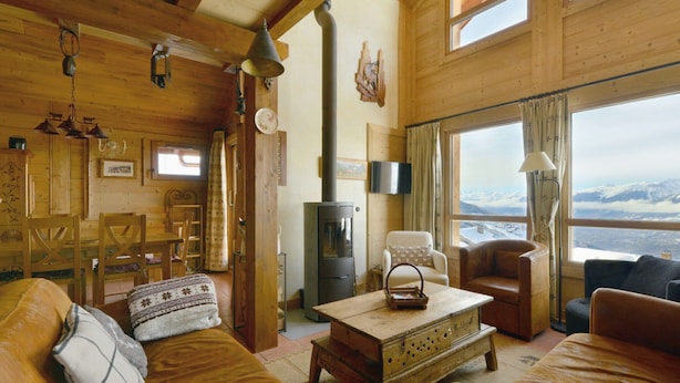 L’intérieur chaleureux d’un chalet avec des murs en bois, une cheminée, un coin repas et une vue sur des montagnes enneigées.