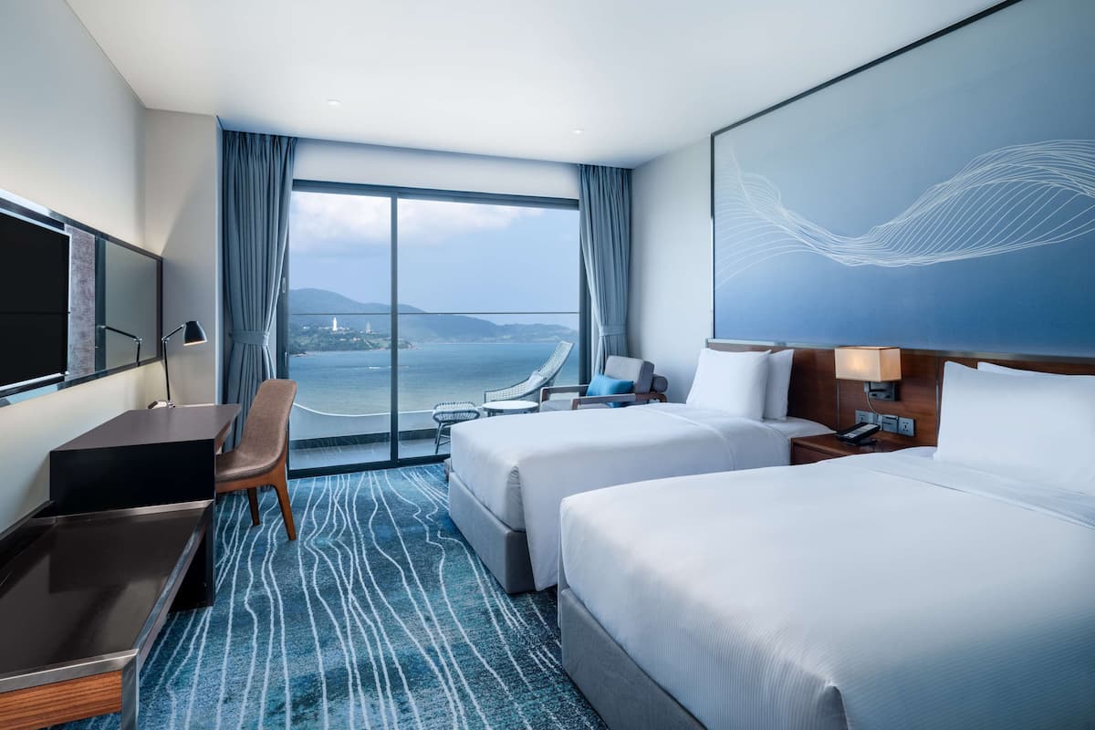 Habitación de hotel con dos camas, un escritorio, una silla, un televisor y un ventanal con vistas al mar.