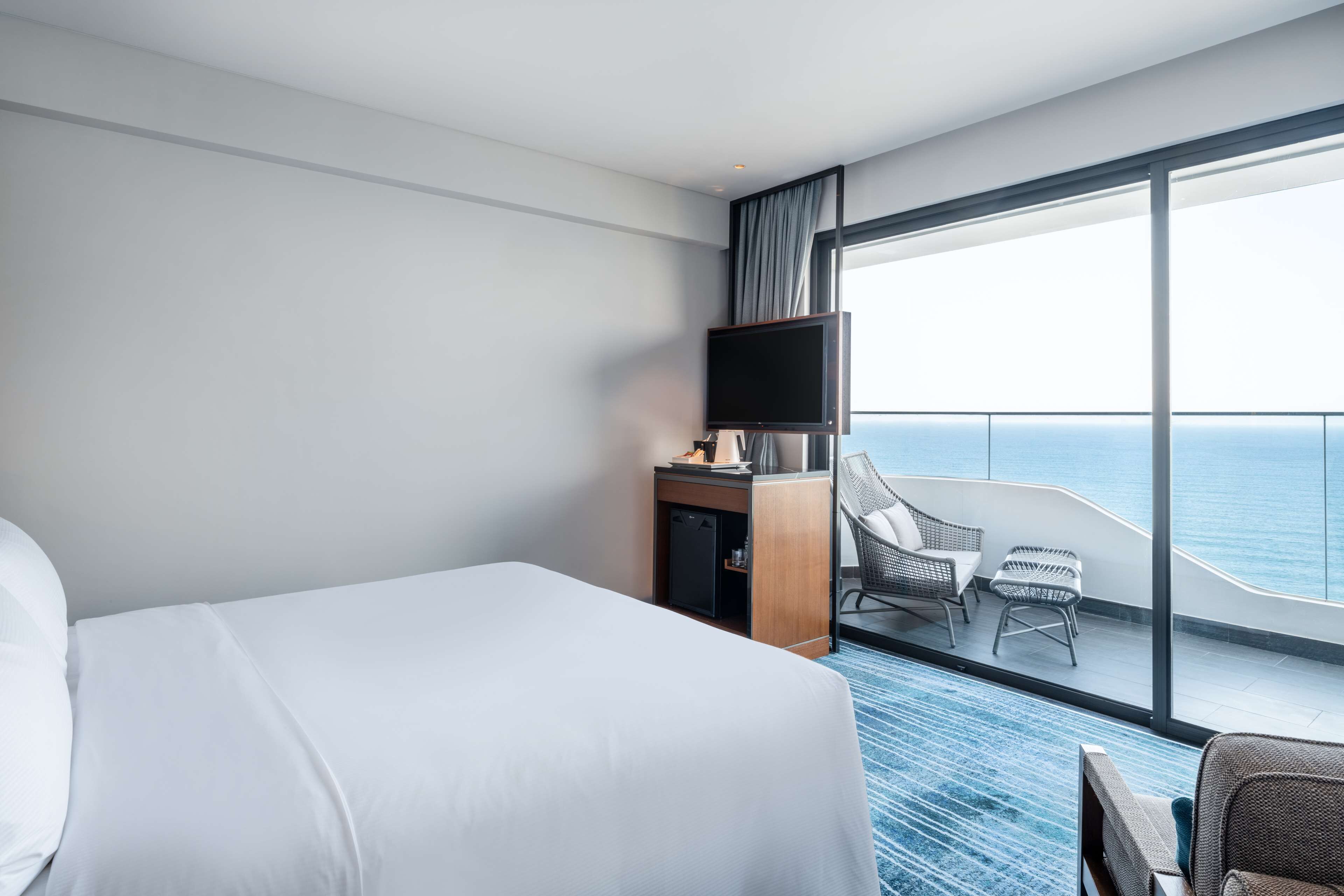 Une chambre d’hôtel avec un grand lit, un bureau avec une télévision et un balcon équipé de chaises, offrant une vue sur l’océan.