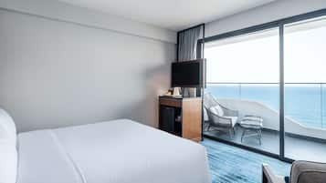 Une chambre d’hôtel avec un grand lit, un bureau avec une télévision et un balcon équipé de chaises, offrant une vue sur l’océan.