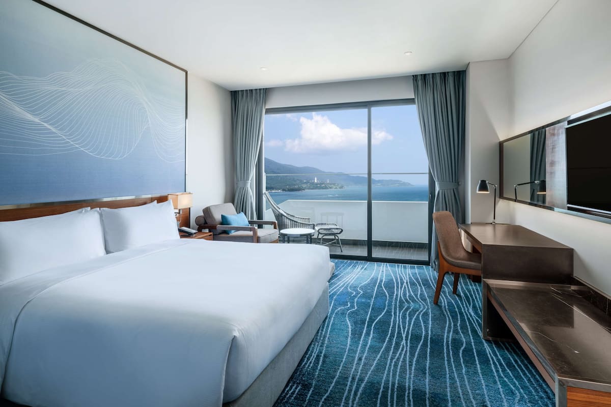 Habitación de hotel con una cama grande, un escritorio, dos sillas, un televisor y vistas al mar y a un puente.