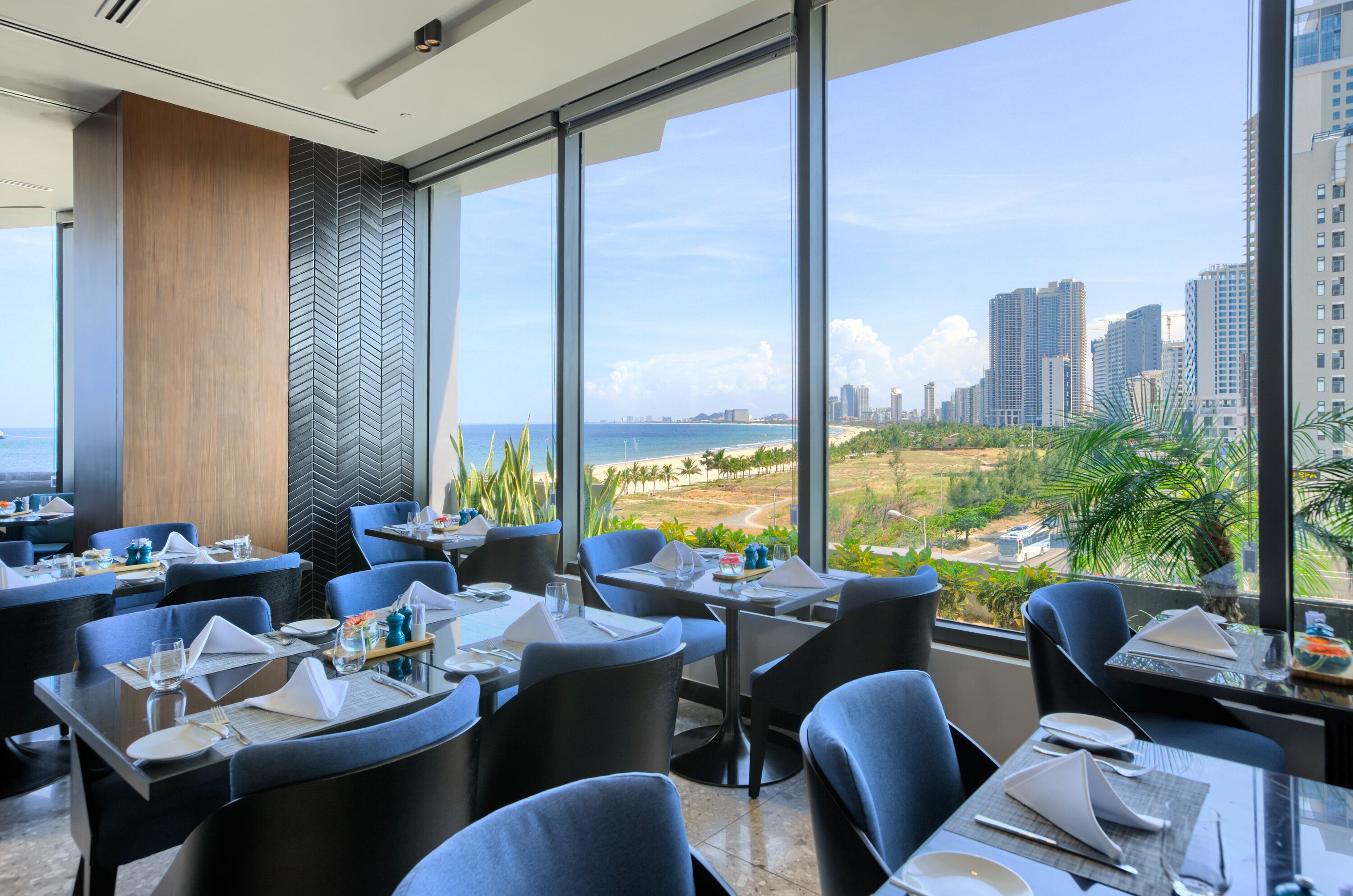 Un espace de restauration moderne, avec des tables dressées pour les clients, de grandes fenêtres offrant une vue sur la ville et la plage.
