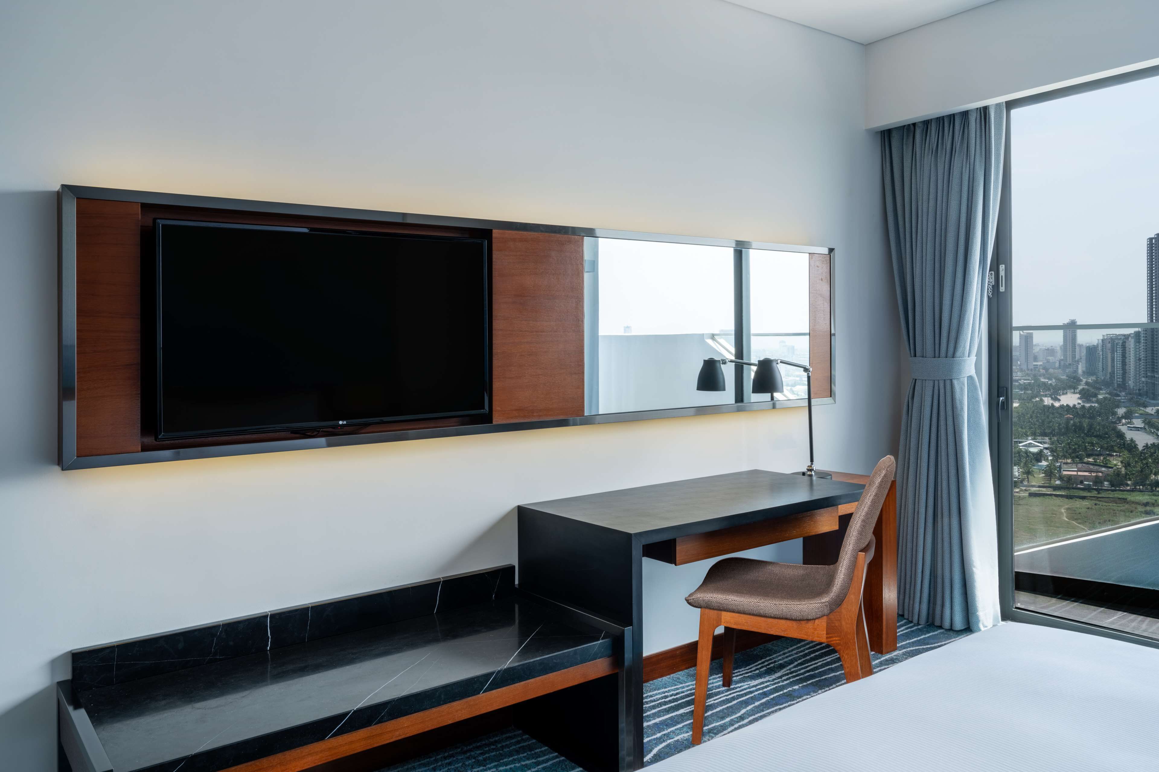 Een moderne hotelkamer met een grote flatscreen televisie aan de muur, een houten bureau met een stoel, en een groot raam met uitzicht over de stad.