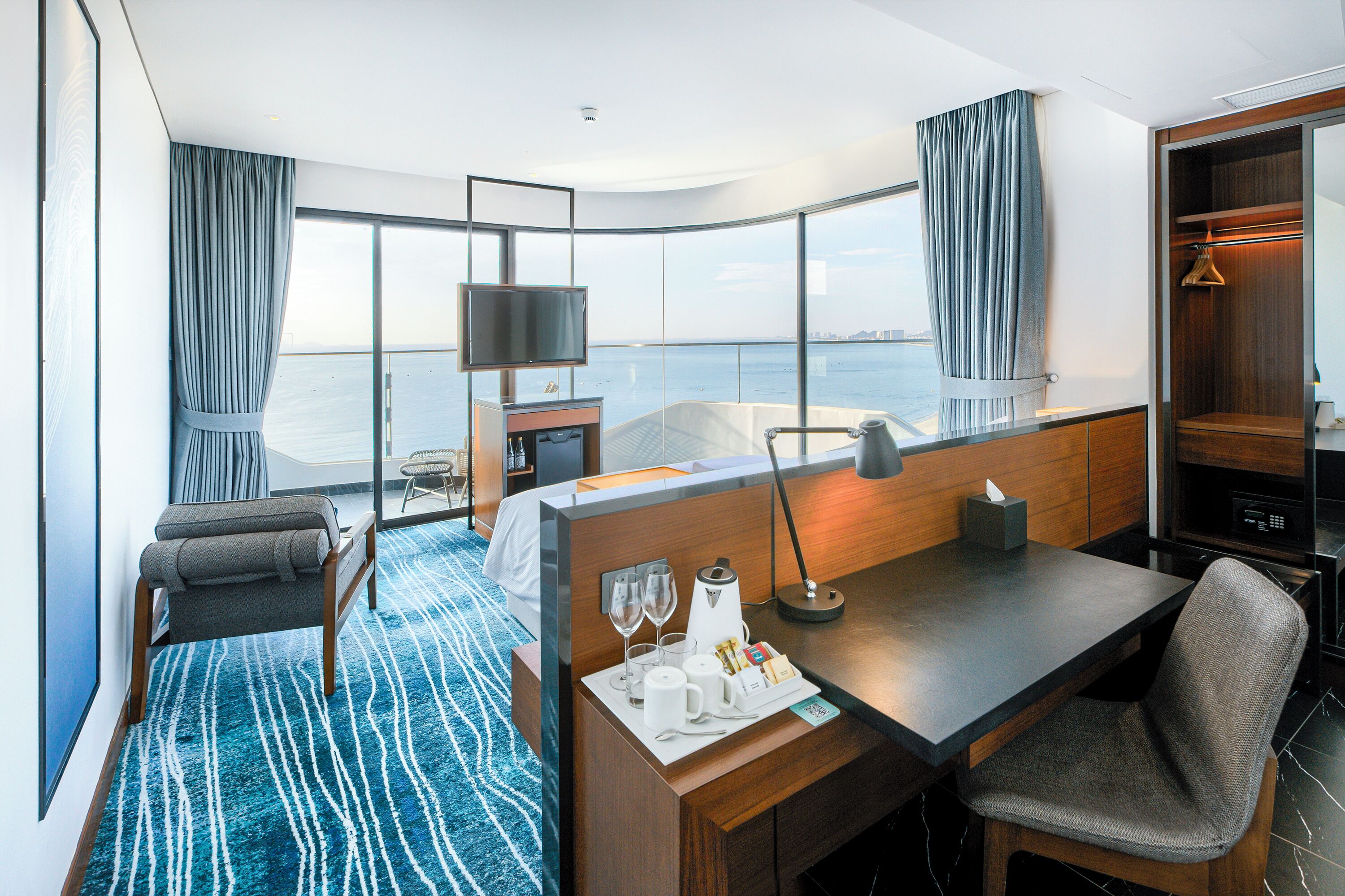 Ein modernes Hotelzimmer mit großem Fenster, Schreibtisch mit Stuhl, Bett, Minibar und Fernseher.