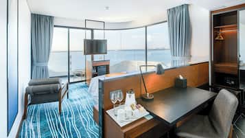 Una habitación de hotel moderna con un ventanal grande, un escritorio con silla, una cama, un minibar y un televisor.