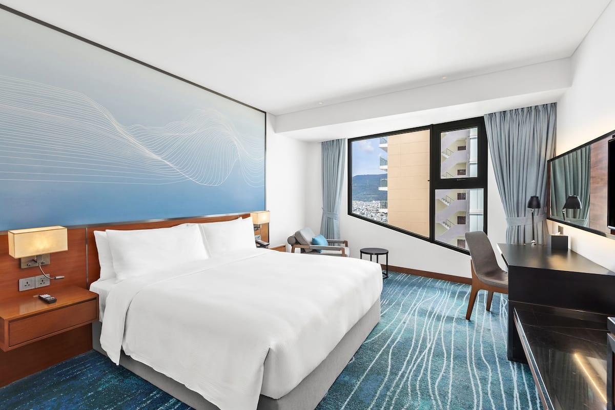 Habitación de hotel con una cama grande, un escritorio, una silla, un televisor y vistas de la ciudad por la ventana.