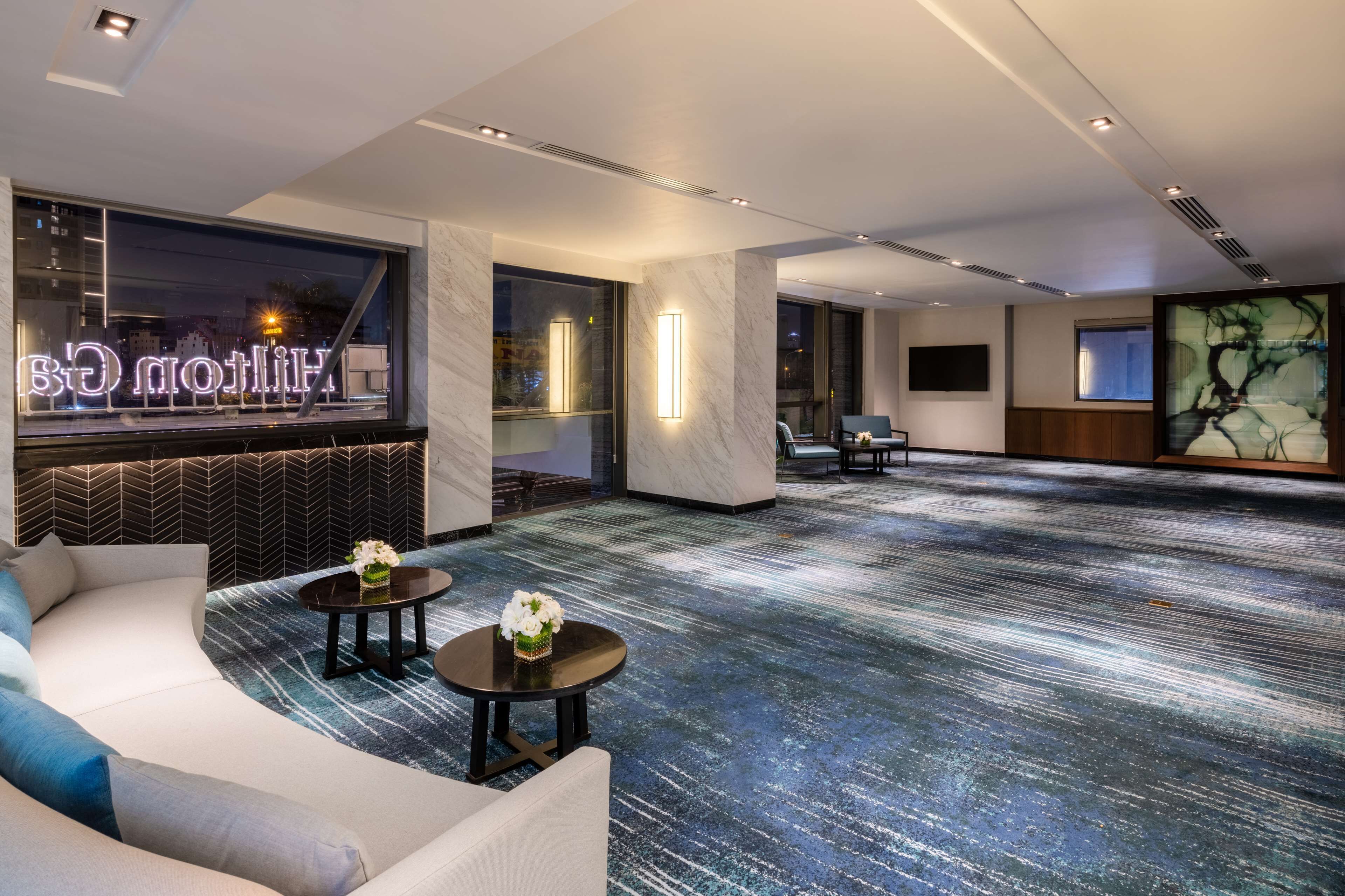 A modern hotel lobby with a curved white sofa, two small tables with floral centerpieces, a patterned carpet, and a large abstract painting on the wall.