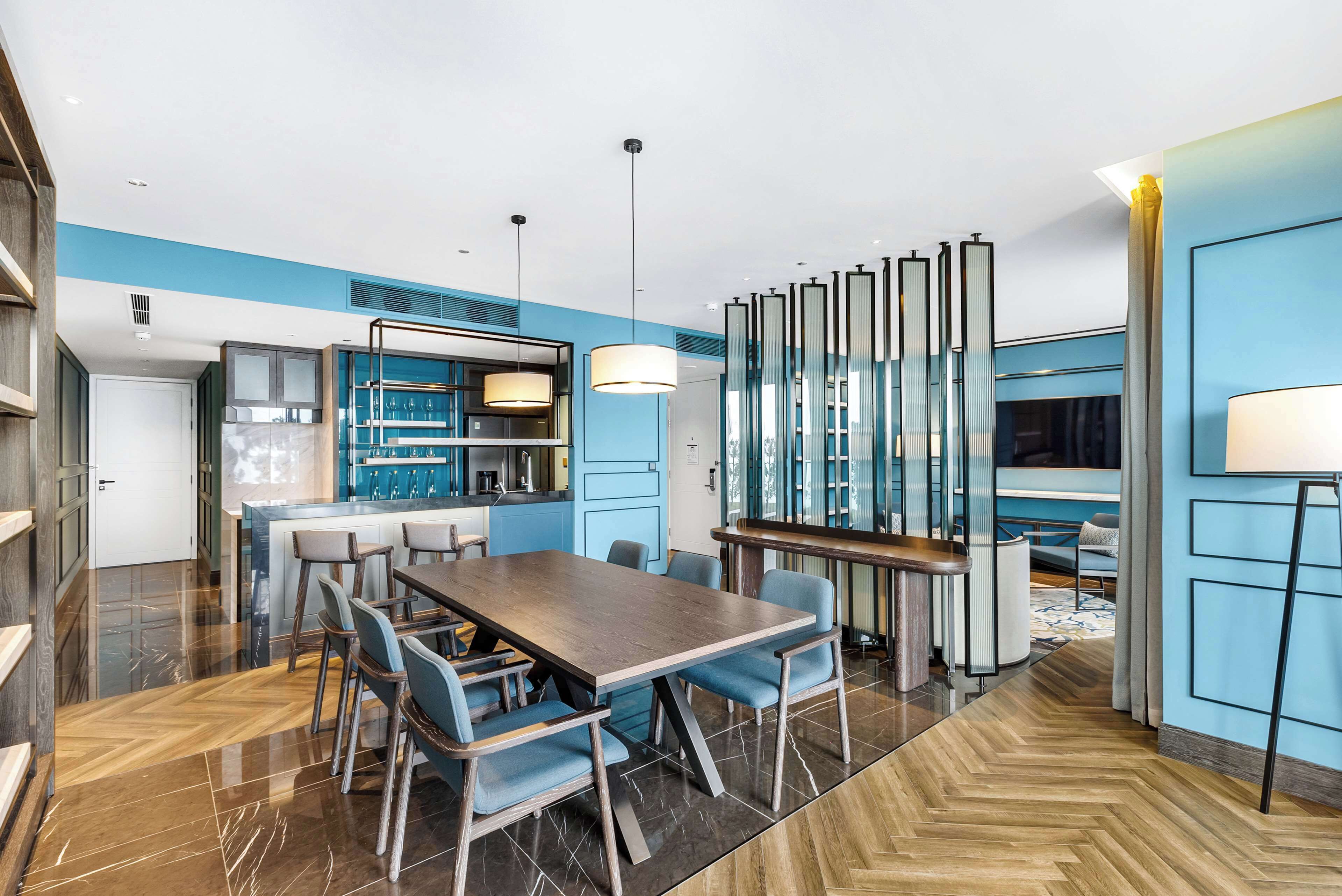 A modern dining area with a long wooden table and blue chairs, a kitchen with blue accents, and a large mirror with vertical panels.