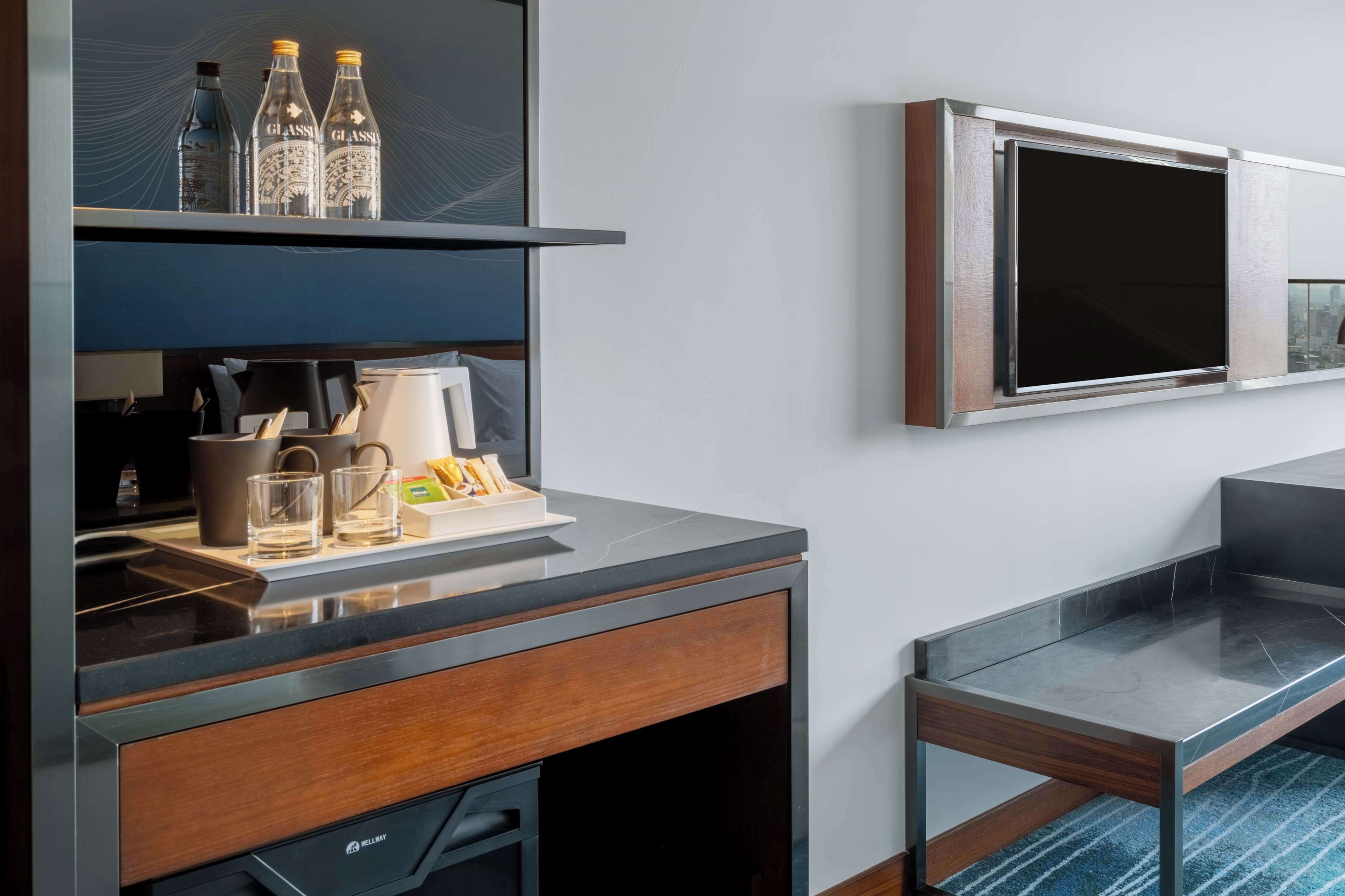A modern hotel room with a built-in kitchen area, a flat-screen TV mounted on the wall, and a small table with a tray of breakfast items.