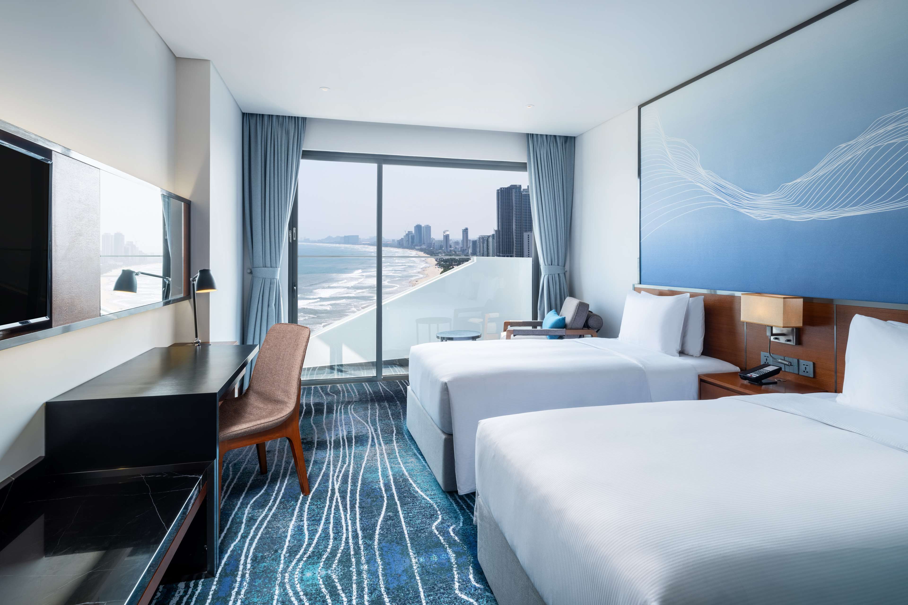 A hotel room with a large window, two beds, a desk, and a chair.