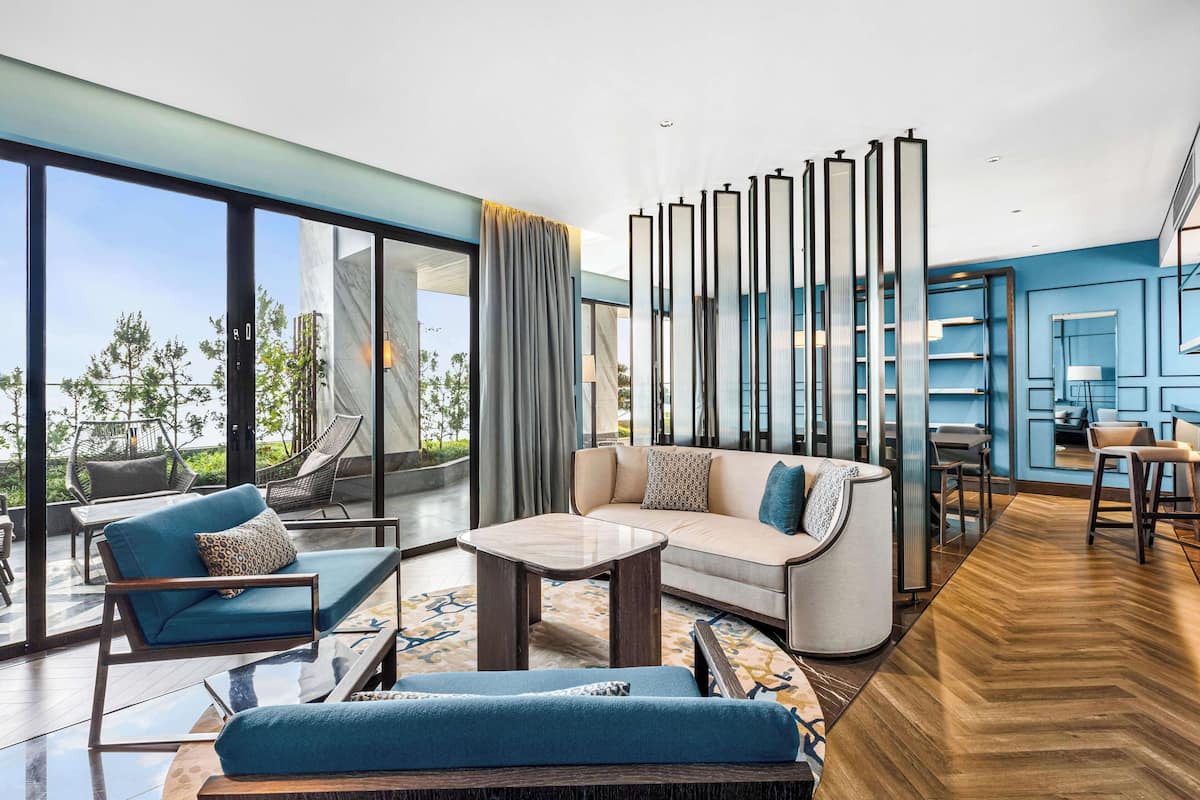 Una sala de estar moderna con sofá, sillones y una mesa de centro. Amplias ventanas ofrecen vistas al exterior.