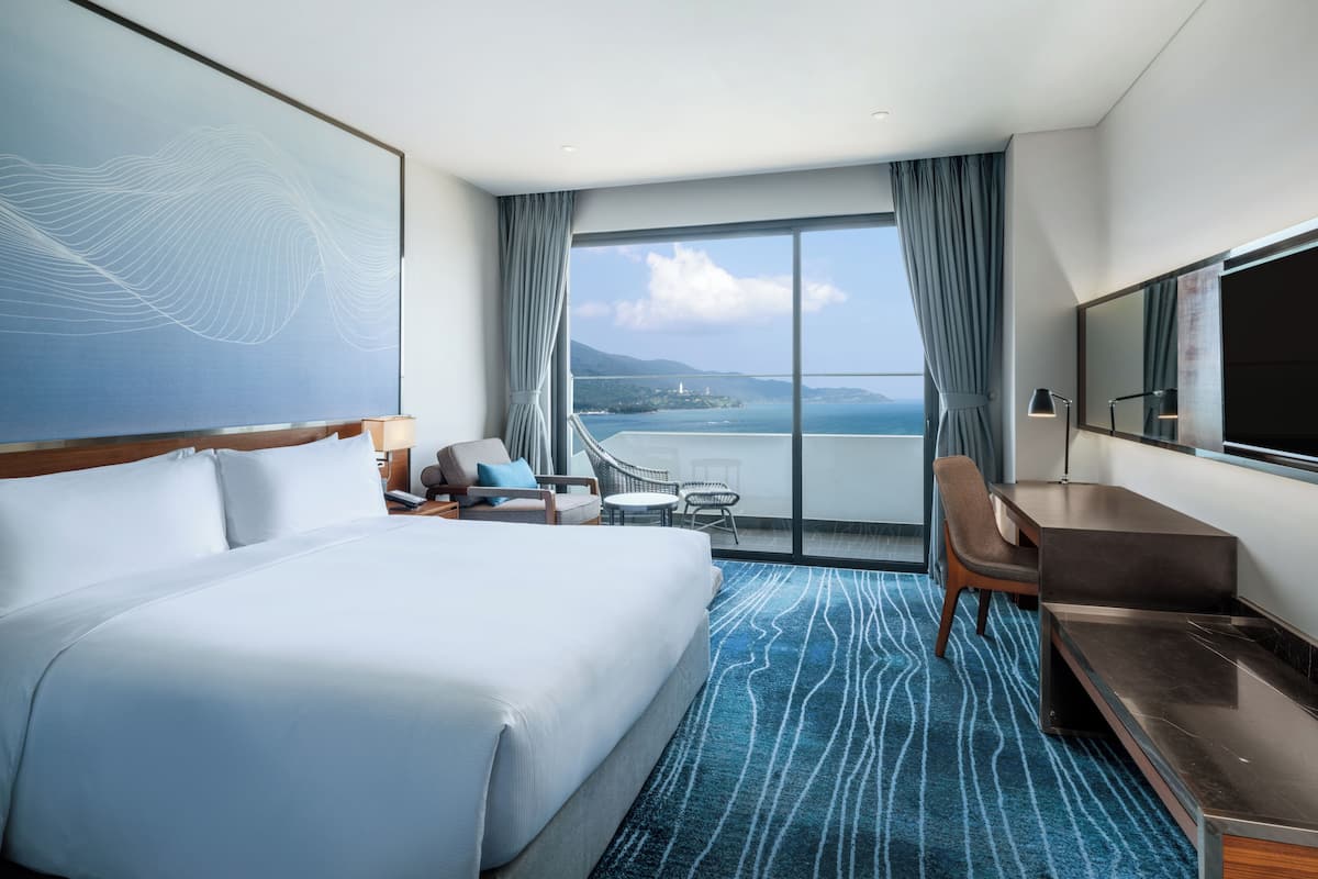 A hotel room with a large bed, a desk, a chair, and a view of the sea.