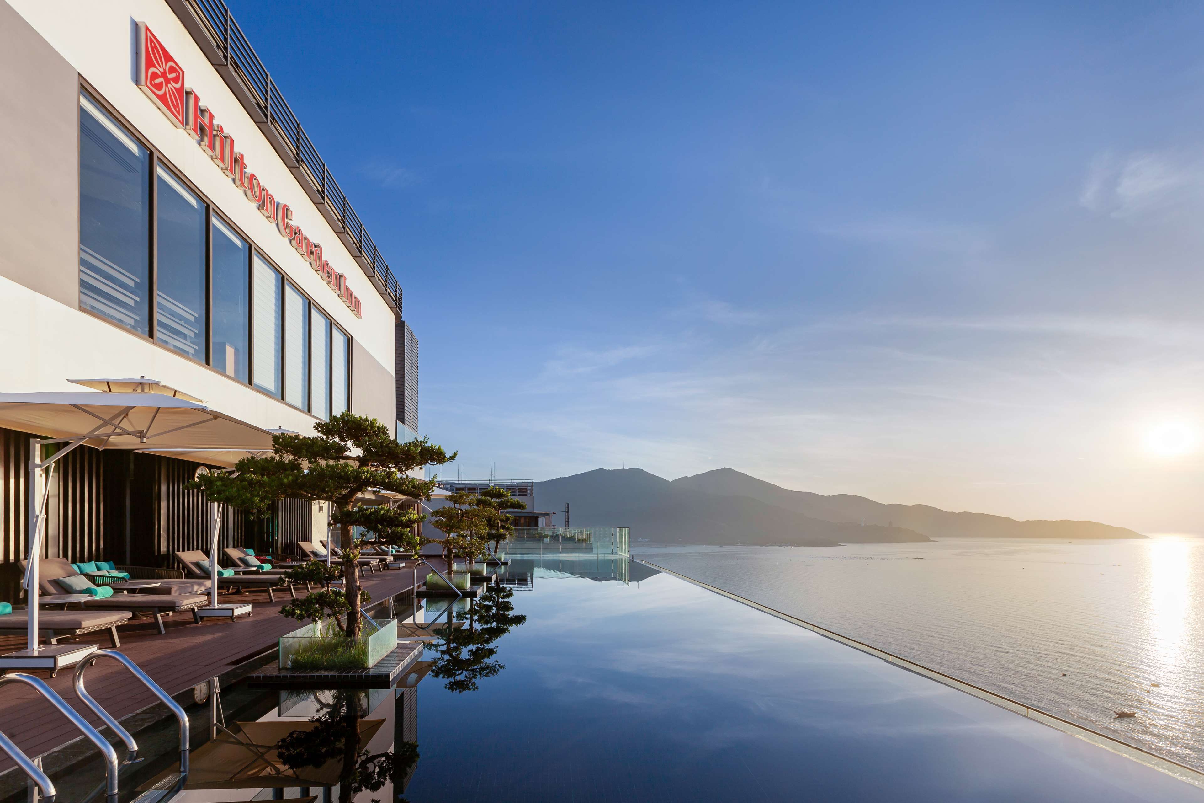 Een hotel met een glazen overloopzwembad, ligbedden en uitzicht op water en bergen.