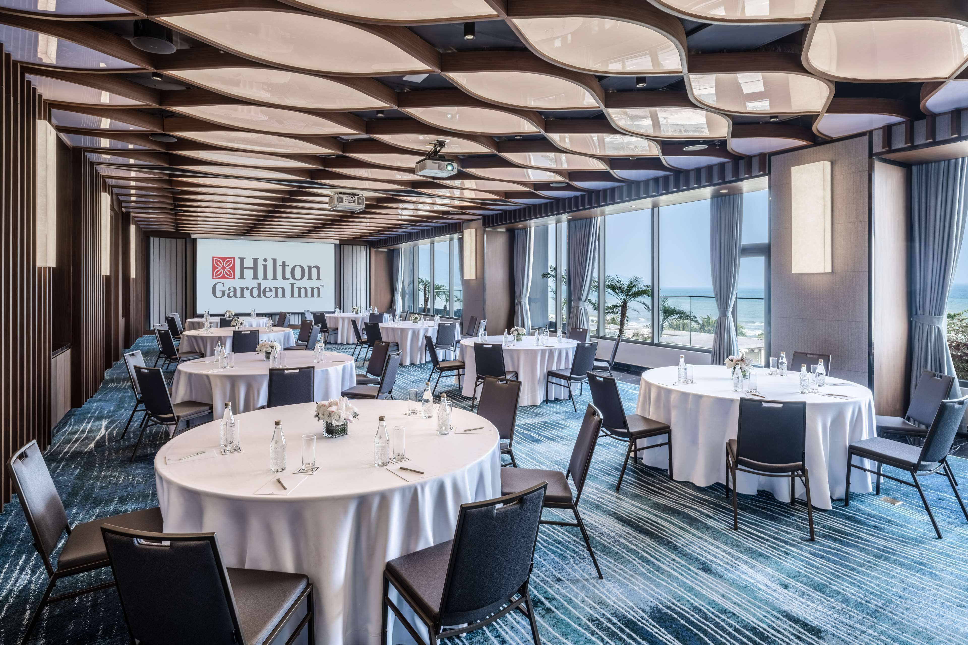 A Hilton Garden Inn conference room with multiple round tables, chairs, and a large screen displaying the hotel's logo.