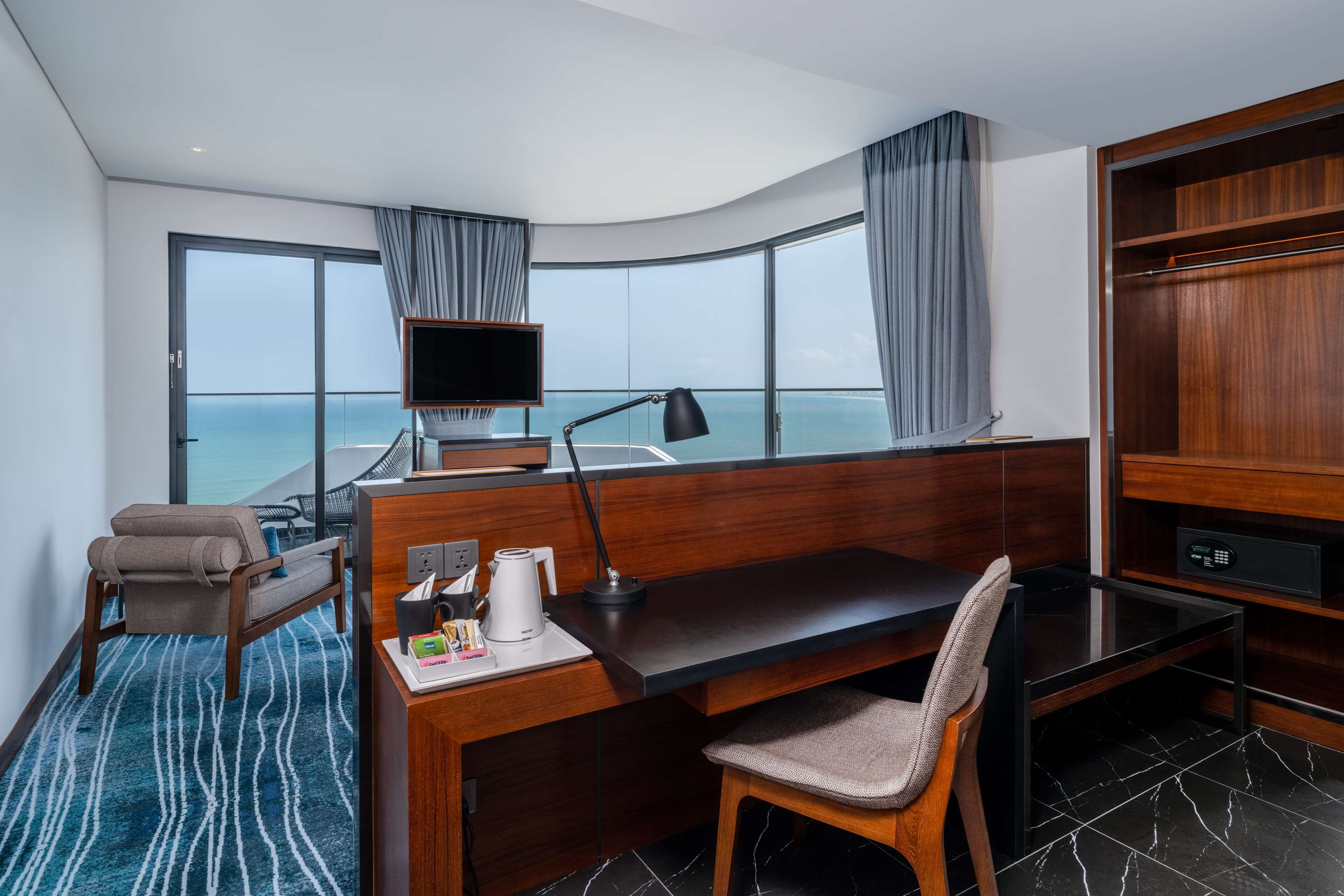 A modern hotel room with a large window offering a view of the sea, a desk with a chair, a television, and a minibar.