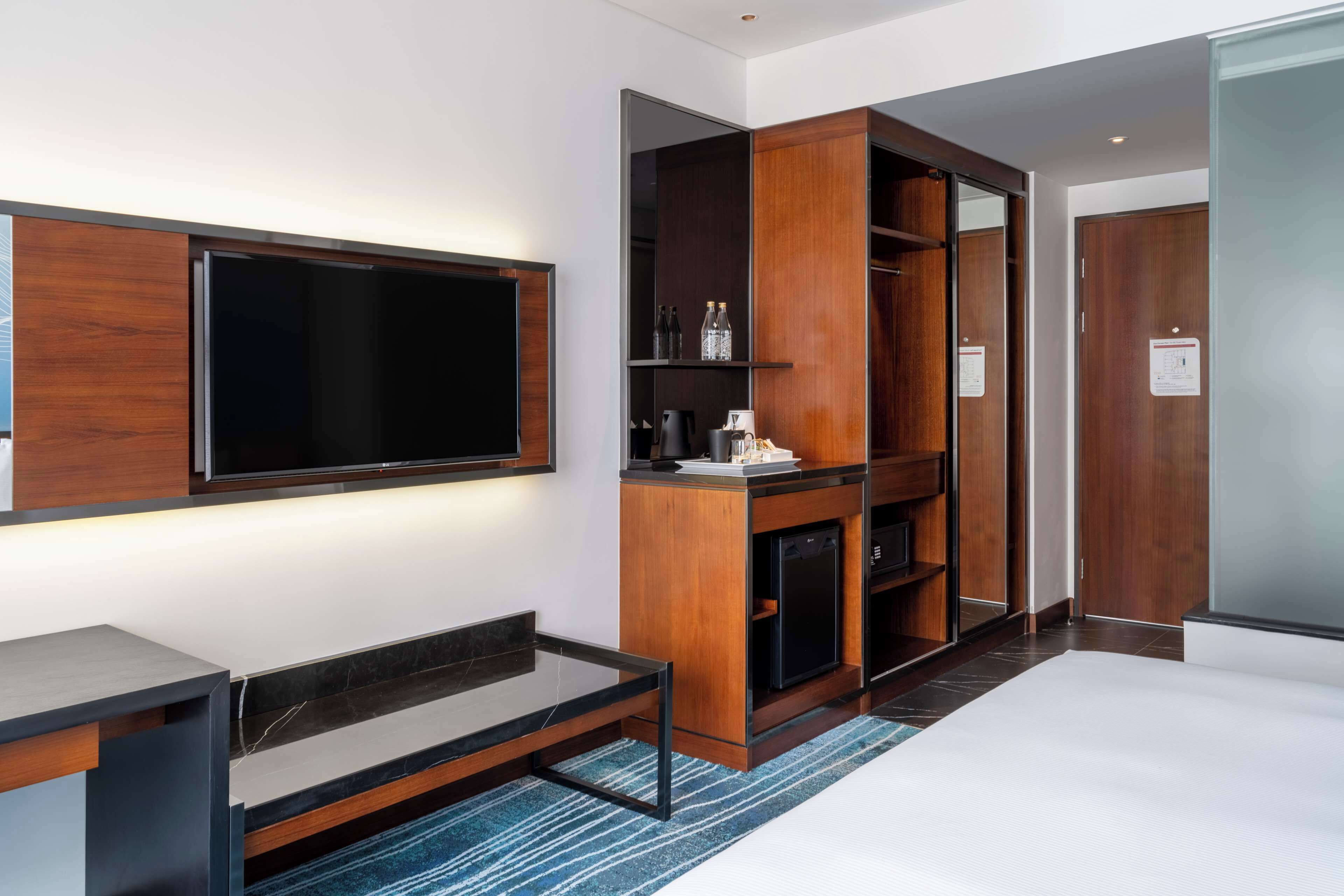 A modern hotel room with a large flat-screen TV, wooden cabinetry, a built-in coffee machine, and a compact refrigerator.