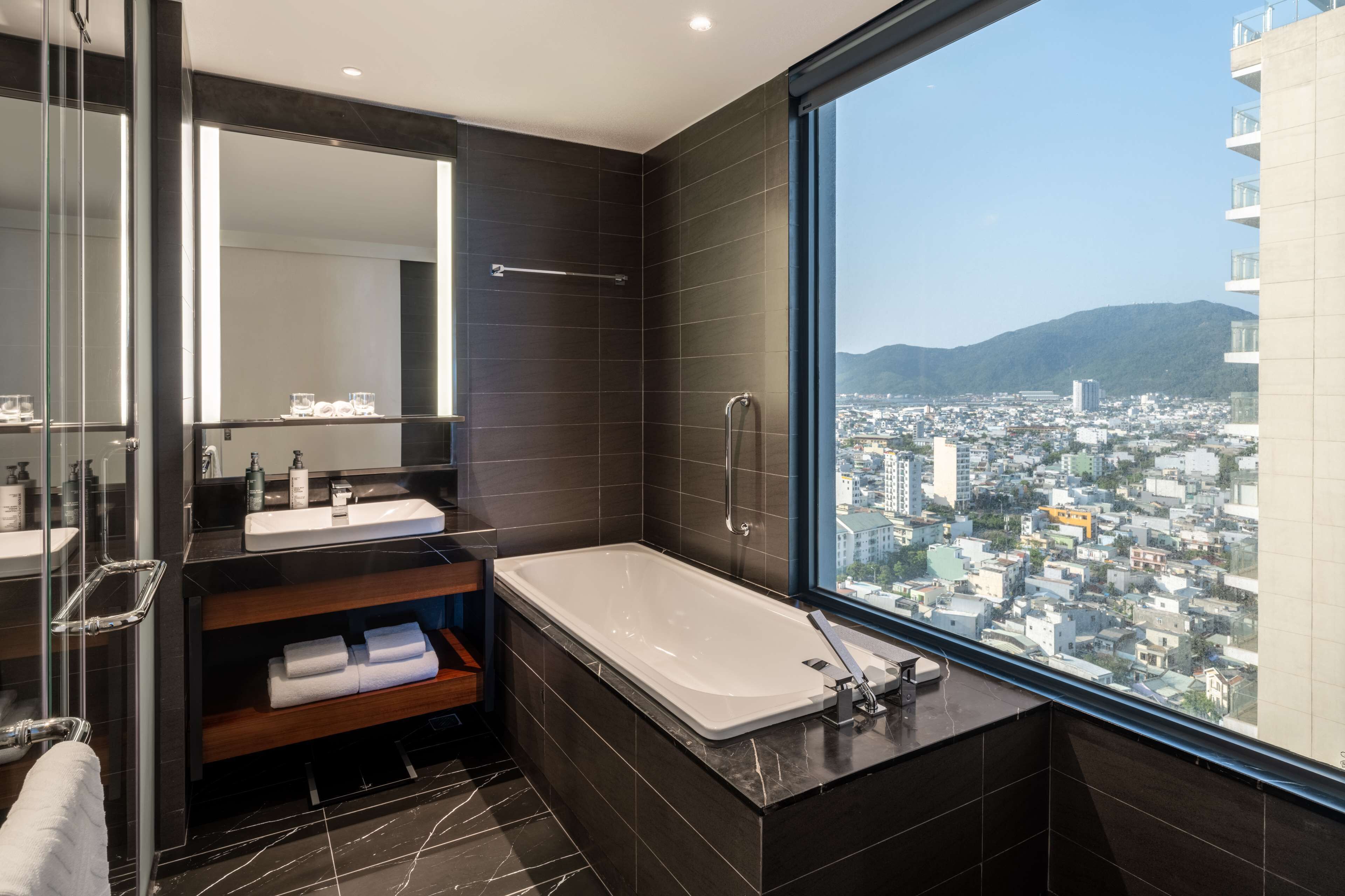 A modern bathroom with a large bathtub, a vanity with a mirror, and a view of a cityscape through a large window.