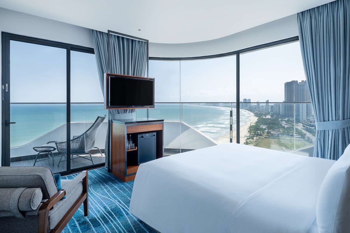 Habitación de hotel con una cama grande, televisión, escritorio y una silla. Tiene vista al océano y al paisaje urbano.