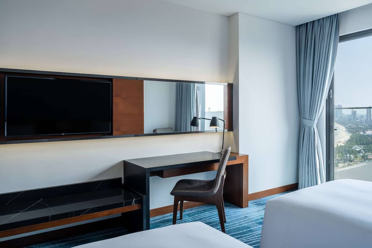 Habitación de hotel con una gran televisión de pantalla plana, un escritorio con silla, un ventanal con cortinas y una cama.