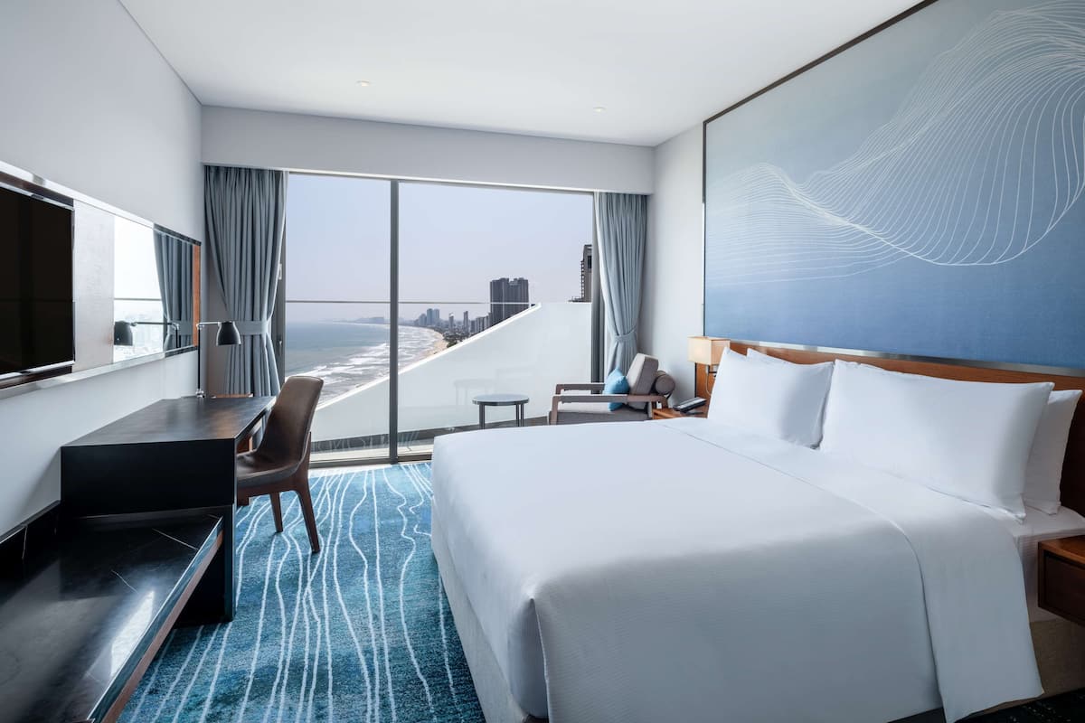 Habitación de hotel con una cama grande, un escritorio, una silla, un televisor y vistas a la playa y al horizonte de la ciudad.
