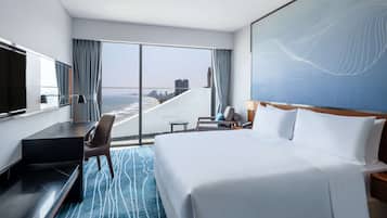 A hotel room with a large bed, a desk, a chair, a TV, and a view of the beach and city skyline.