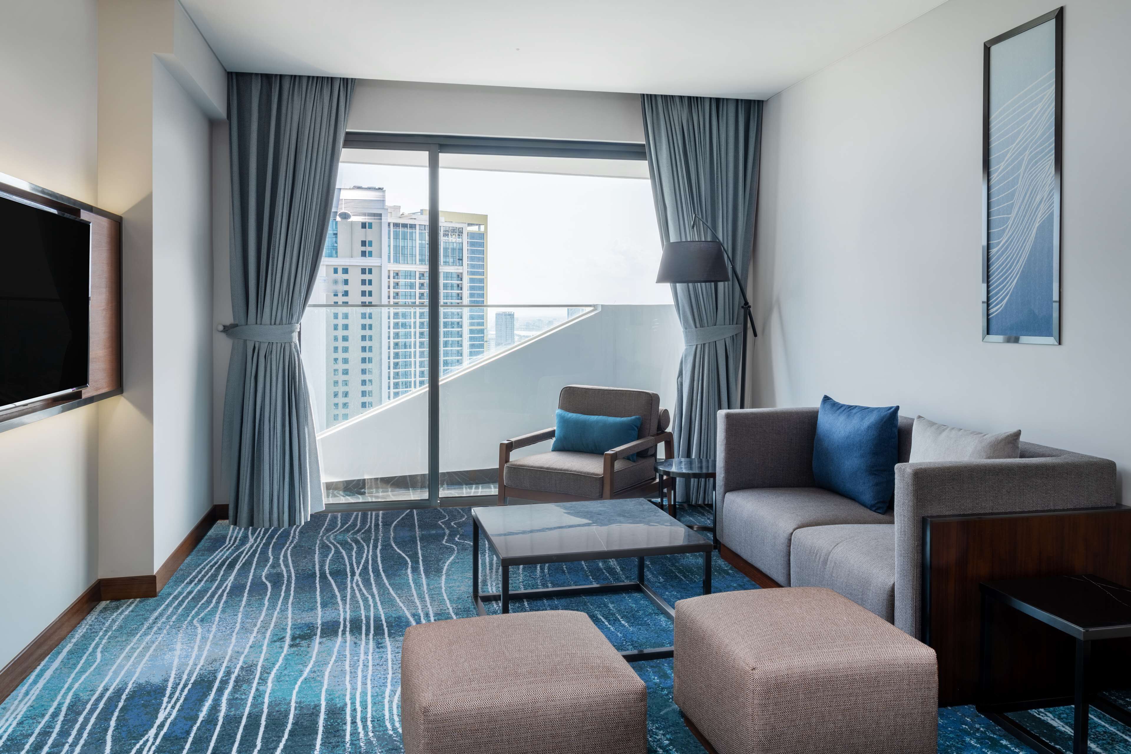 A modern hotel room with a large window, a flat-screen TV, a sofa, a chair, and a coffee table.
