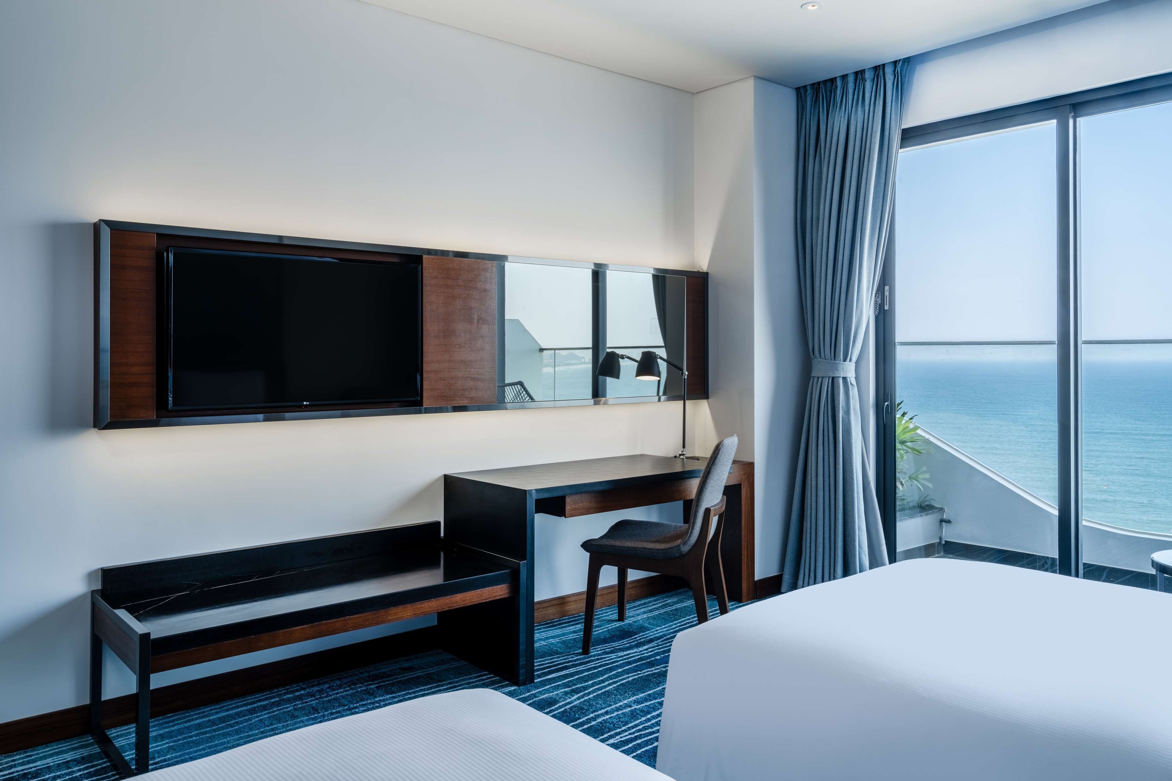 Ein Hotelzimmer mit Bett, Schreibtisch, Stuhl, Fernseher und einem großen Fenster mit Meerblick.