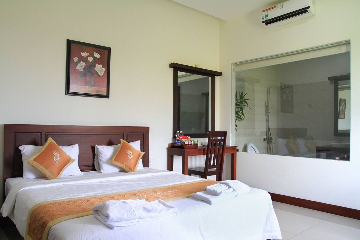 A hotel room with a bed, a desk, a chair, a mirror, and a bathroom visible through a glass door.