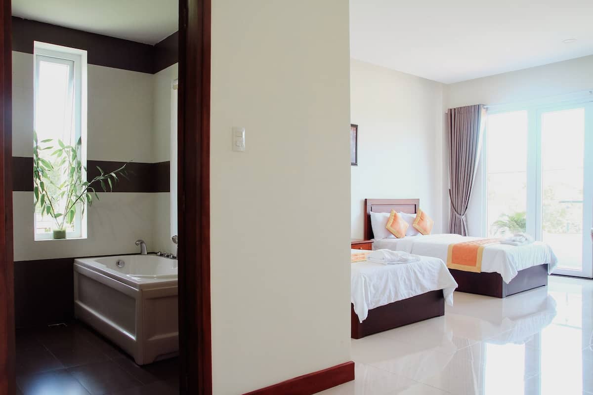 A hotel room with a bathtub, two beds, a window with curtains, and a view of greenery outside.