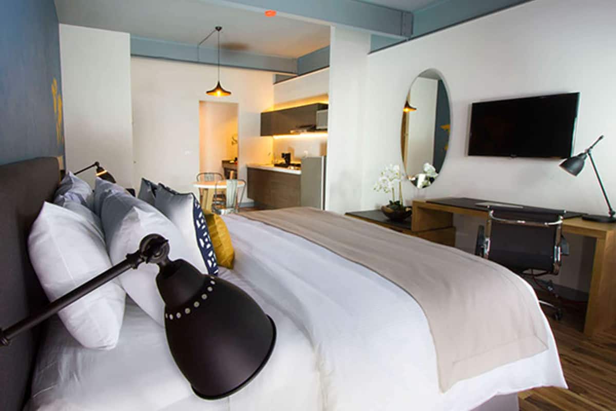 Une chambre d’hôtel avec un lit, un bureau, une chaise, une télévision et, au fond, un coin cuisine.