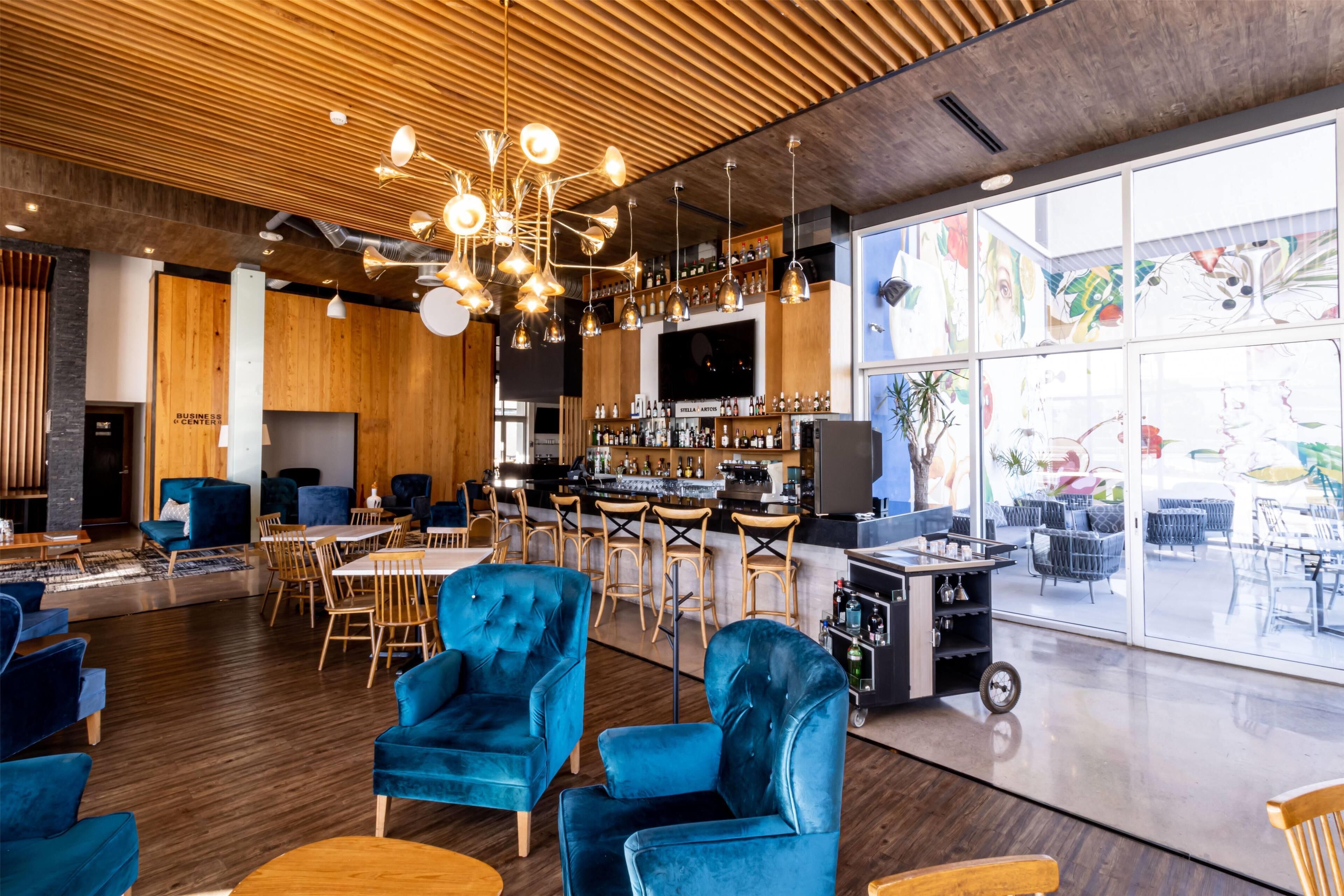 A modern bar area with wooden ceiling, blue velvet chairs, and a bar counter with stools.