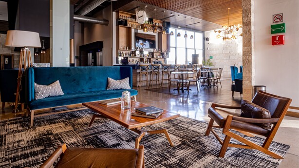 A modern lounge area with a blue sofa, wooden coffee table, and leather armchairs. There is a bar area in the background with shelves of bottles and a television. - TRYP by Wyndham San Luis Potosi Hotel & Suites (San Luis Potosi)