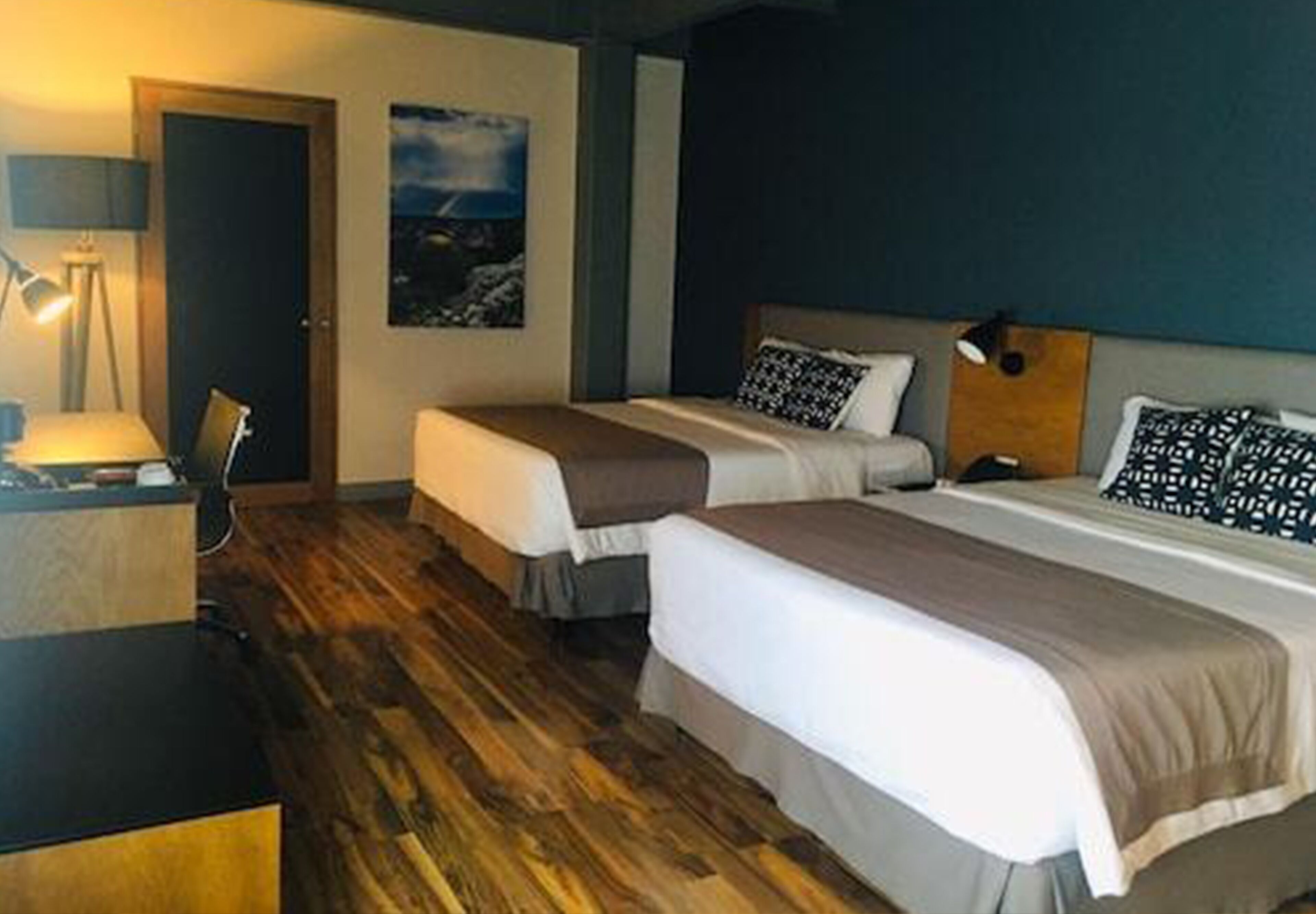 A hotel room with two beds, wooden flooring, a desk, and a lamp.
