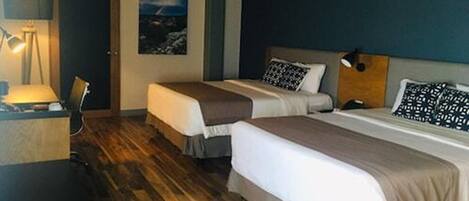 A hotel room with two beds, wooden flooring, a desk, and a lamp.