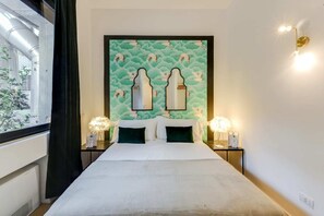 Une chambre d’hôtel avec un grand lit, deux tables de chevet avec des lampes, un mur orné d’une fresque représentant des nuages et des oiseaux, et une fenêtre donnant sur un paysage verdoyant.