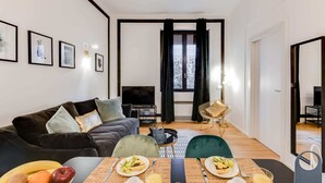 Uma sala de estar moderna com um sofá, uma mesa de jantar posta com frutas e bebidas, uma televisão e uma janela com cortinas.