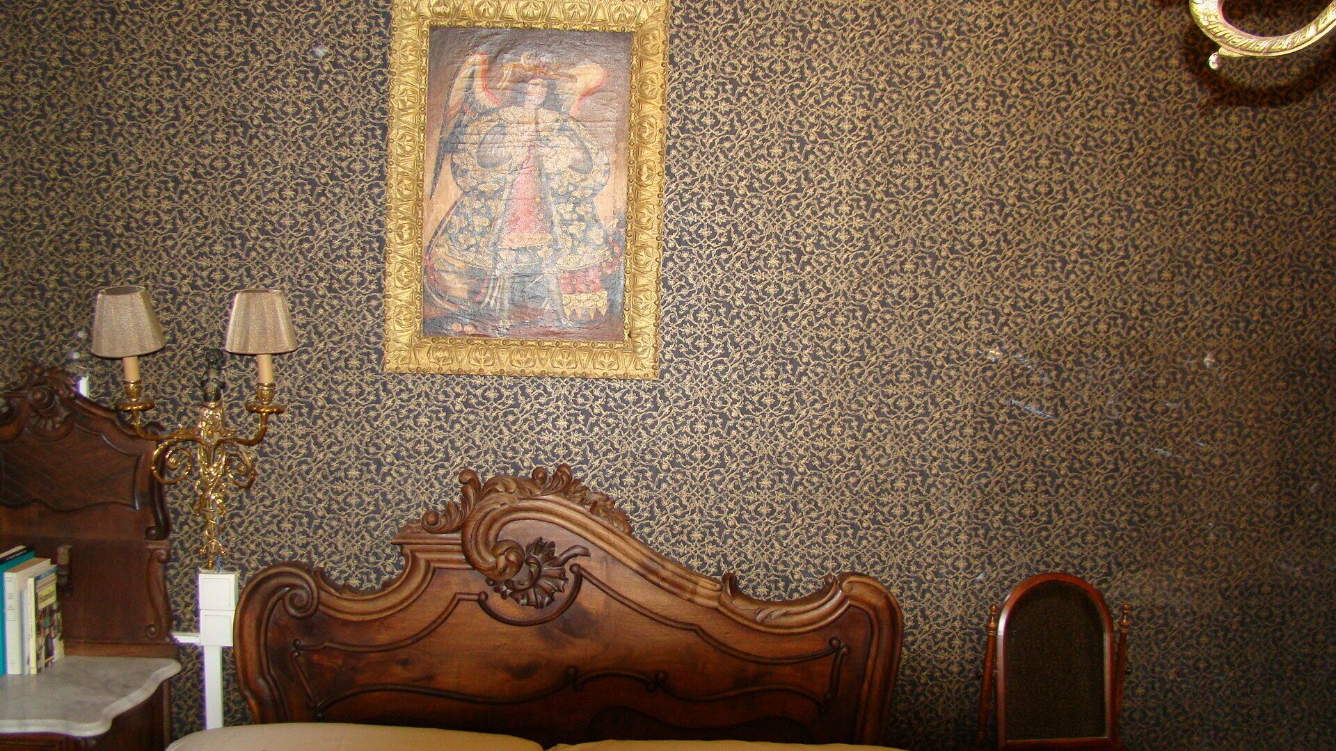 Een slaapkamer met behang met een patroon, een houten bed met een versierd hoofdeinde, een nachtkastje met lampen, en een ingelijst schilderij aan de muur.