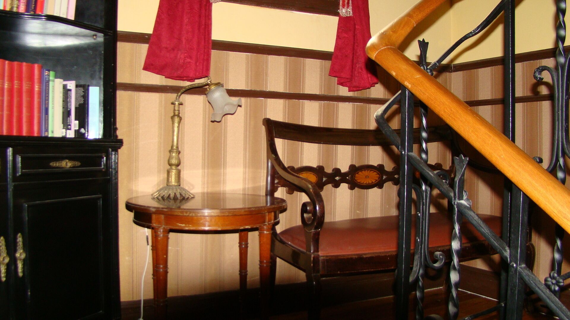 A wooden chair with a cushioned seat and backrest, a small wooden table with a lamp, and a bookshelf with books.