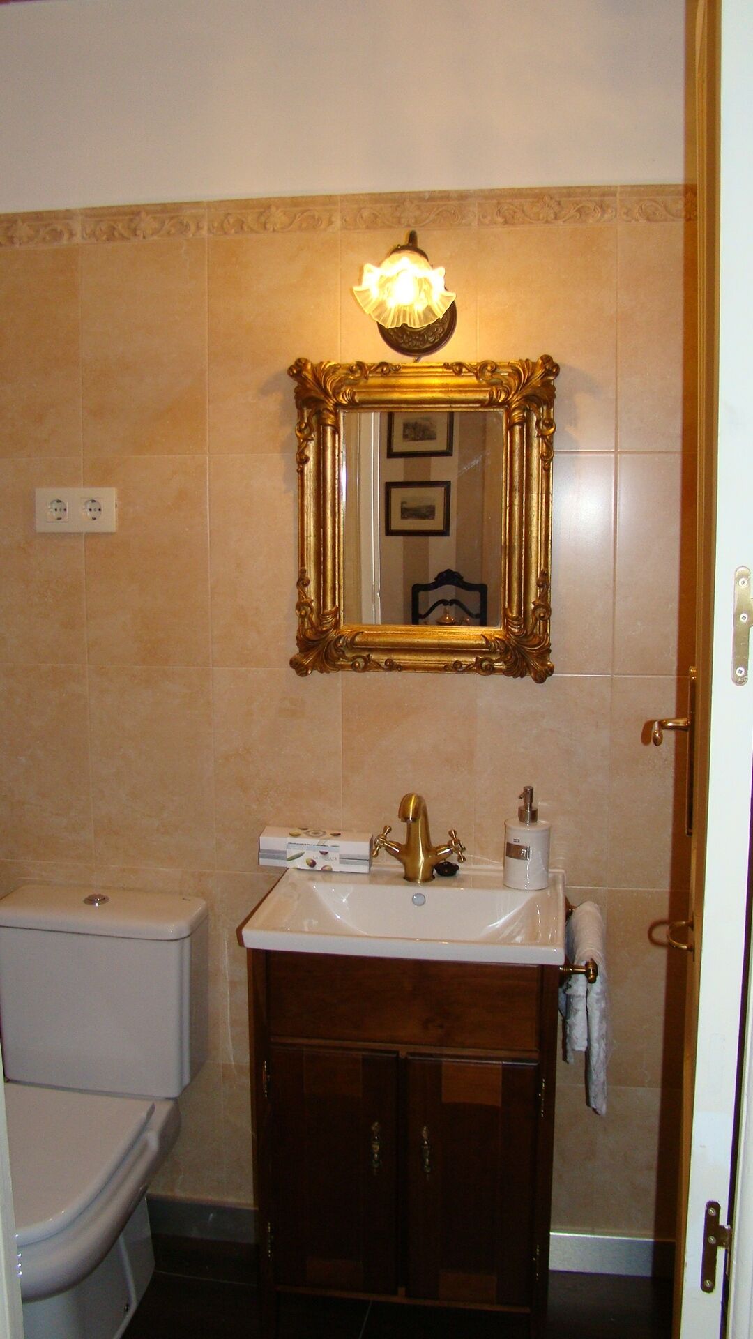 Een badkamer met een spiegel met gouden lijst, een wit wastafelmeubel met gouden kraan, en een wit toilet.