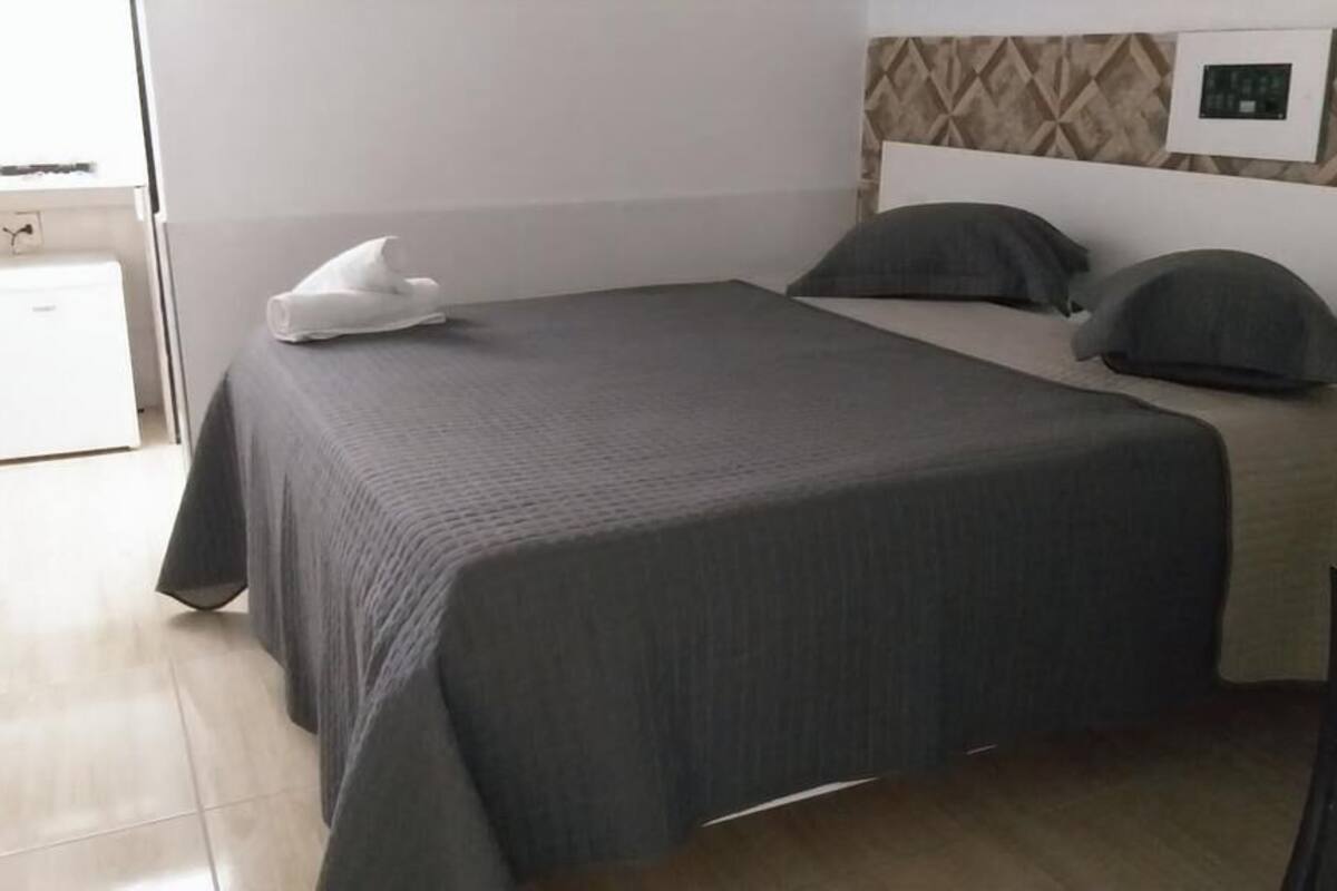 Habitación de hotel con una cama grande, un refrigerador blanco pequeño y una ventana con vistas al exterior.