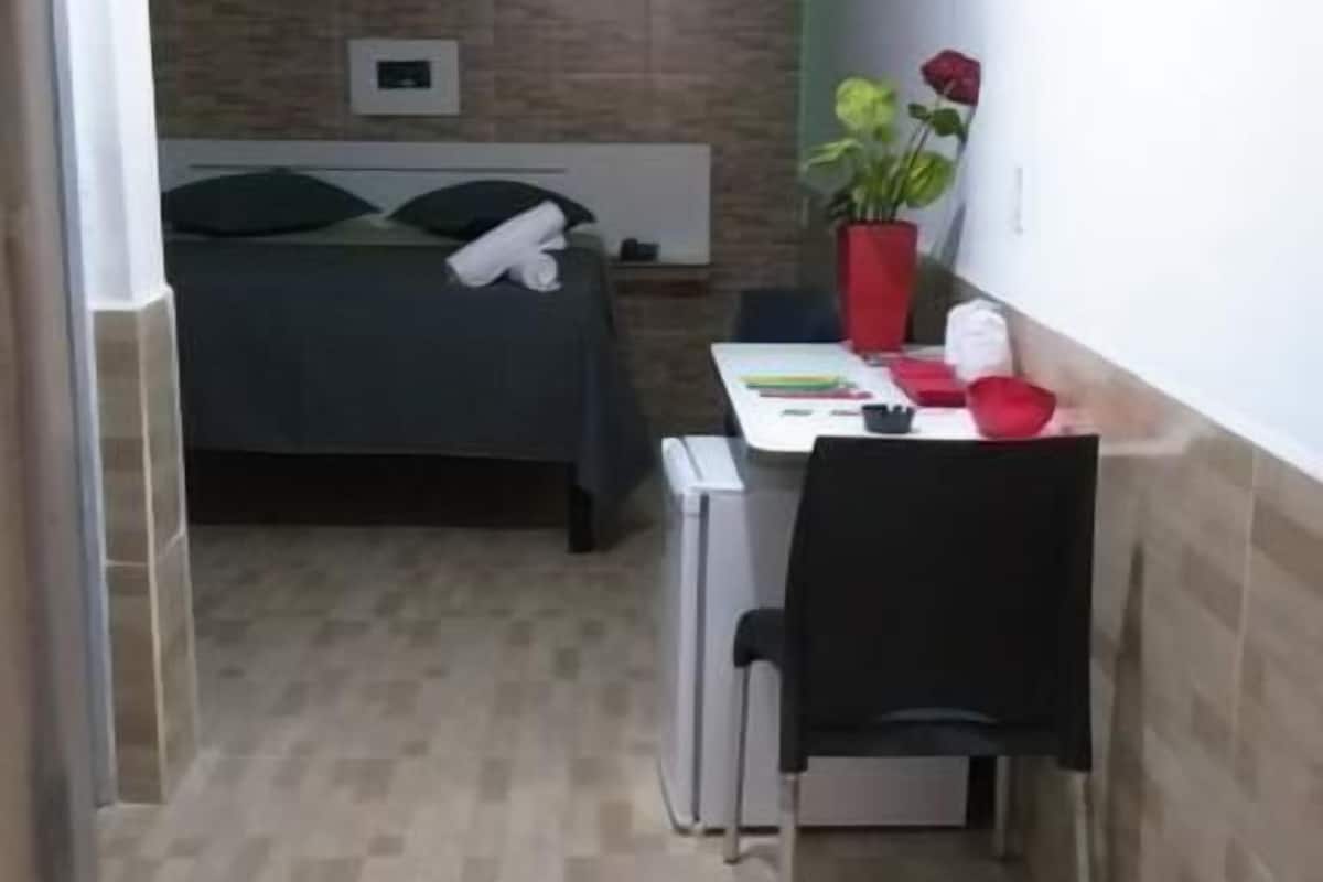 Una habitación pequeña y funcional con una cama, un escritorio y una silla. El piso es de baldosas y hay una tira de luz verde en el techo.