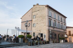 A building with a stone facade and multiple windows, a cobblestone street, outdoor seating with tables and chairs, and a sign for a hotel. - Hotel Panorama (Bertinoro)