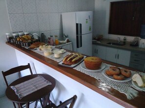 Una cocina con mostrador que exhibe varios productos horneados, un refrigerador y un comedor con una silla y una mesa.