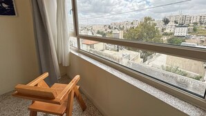 A room with a large window, a wooden chair, and a view of a cityscape. - Carob Hostel (Amman)