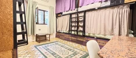 Ein Zimmer mit Etagenbetten, einem kleinen Tisch, einem Stuhl, einem gemusterten Teppich und einem Fenster mit Vorhängen.