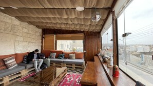 A cozy room with a wooden ceiling, a sofa with cushions, a wooden coffee table, and a view of the city through large windows. - Carob Hostel (Amman)