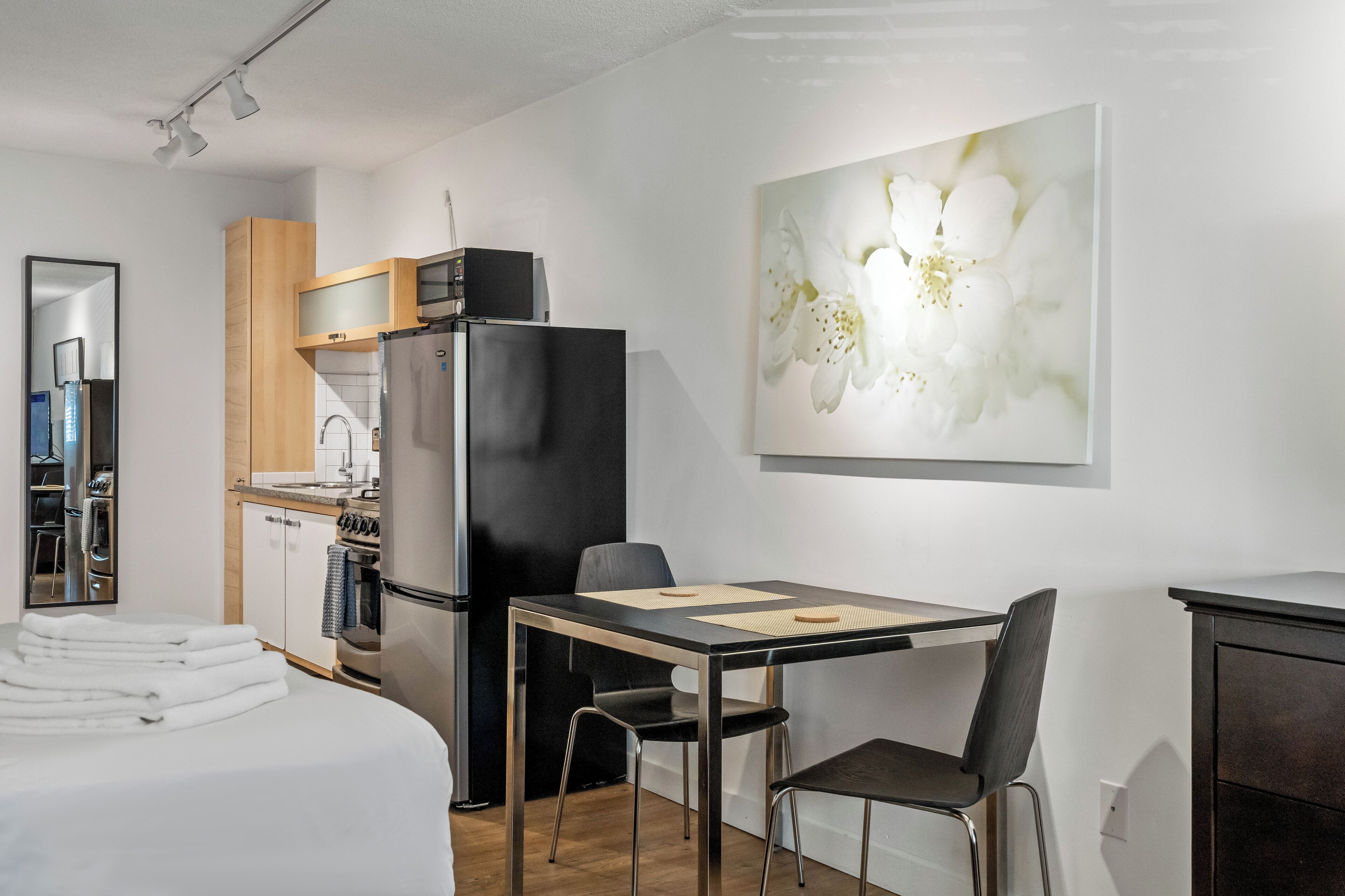Ein modernes Hotelzimmer mit einem Bett, einem Esstisch mit Stühlen, einer Küchenzeile inklusive Kühlschrank und einem großen Blumenbild an der Wand.