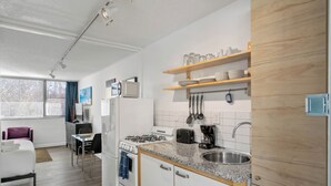 A modern kitchen with white cabinets, granite countertops, and wooden shelves. There is a dining area with a purple chair and a small table.