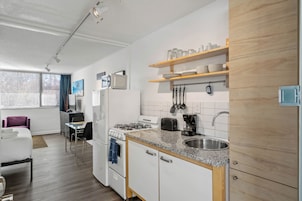 Una cocina moderna con armarios blancos, encimeras de granito y estantes de madera. Hay un rincón comedor con una silla morada y una mesa pequeña.